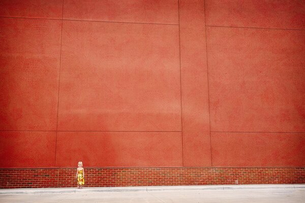 The girl is standing near the red wall on the street