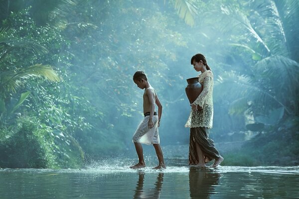A girl with a jug and a boy walk along a river in the jungle