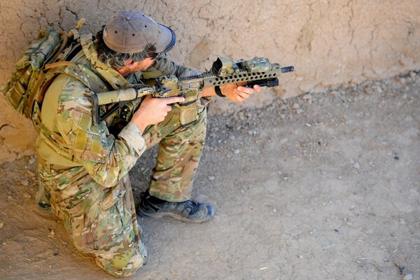 A soldier in camouflage crouches down and takes aim with a rifle